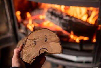 Rise In Price Of Firewood For Heating Concept. Hand Holds Log With Sad Smiley Against Background Of Burning Fireplace.