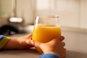 Child's Hands Grabbing A Crystal Glass With Orange Juice On The Kitchen Counter.