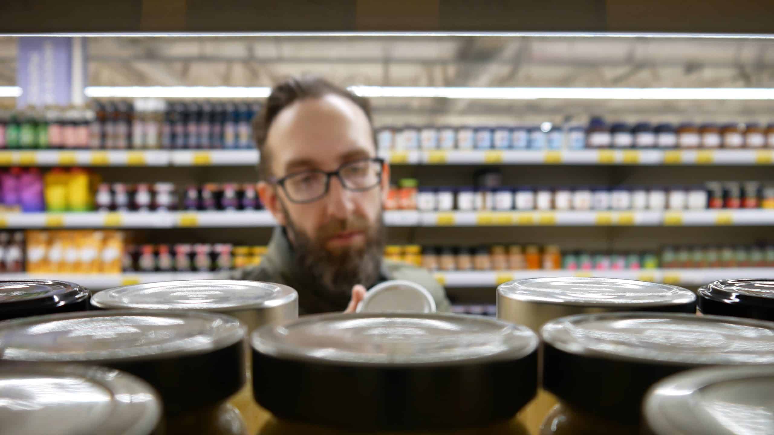 Close Up Of Many Jam Jars With Metallic Lids On A Supermarket Shelf And A Male Buyer With Glasses Choosing One