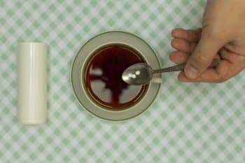 Pour Their Teaspoon Into Tea With Three Small White Sugar Substitute Tablets And A Pill Storage Container Nearby. One Tablet Replaces 1 Teaspoon Of Sugar