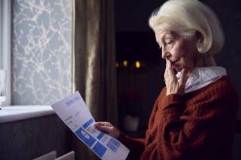 Senior Woman With Usa Energy Bill Trying To Keep Warm By Radiator In Cost Of Living Energy Crisis