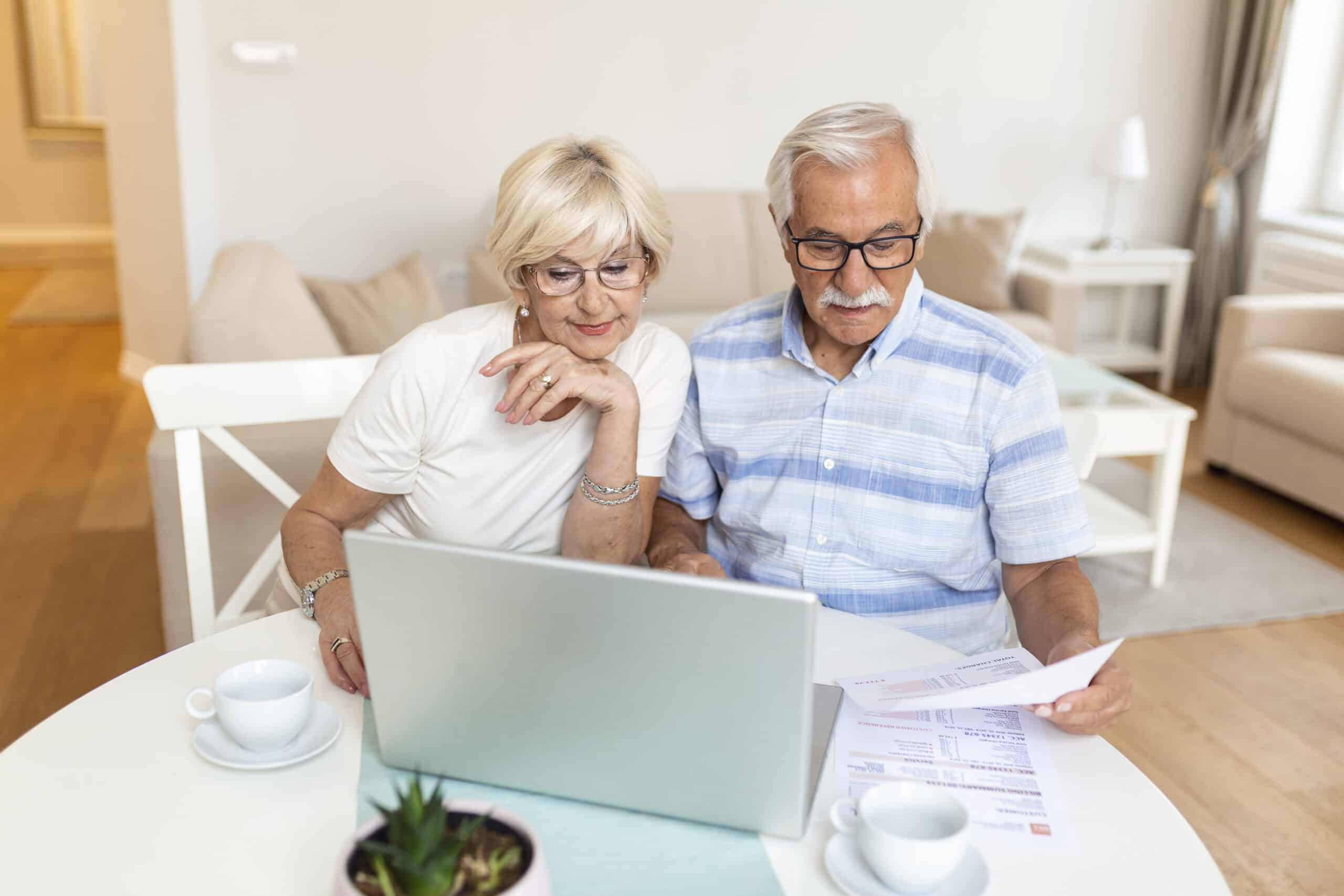 Happy Middle Aged Husband And Wife Sitting At Table With Laptop And Paper Bills, Calculating Domestic Incomes Together At Home.