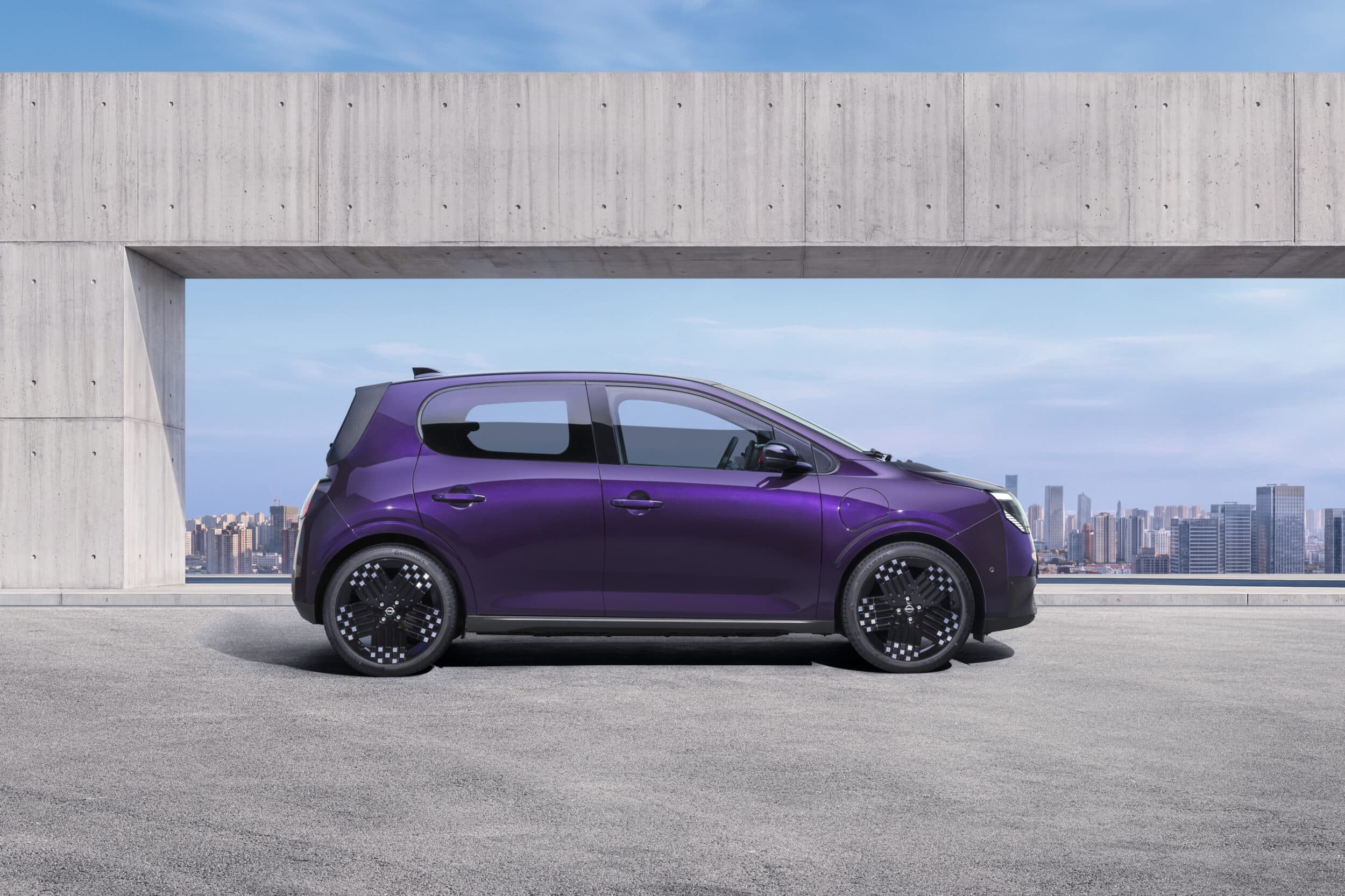 Photograph showcasing a purple Nissan PIXO parked on a rooftop terrace. Side profile highlights aerodynamic shape, black alloy wheels concrete architectural frame, and city skyline backdrop.